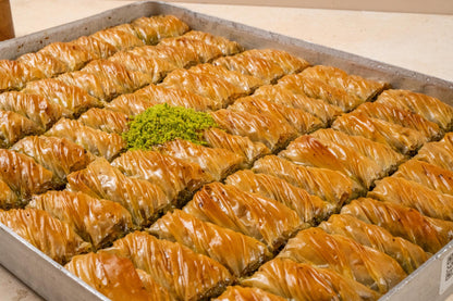 May include: A tray filled with golden-brown baklava pastries, arranged in neat rows. A small pile of green, finely ground pistachios sits in the center. The dessert has a flaky, layered appearance.