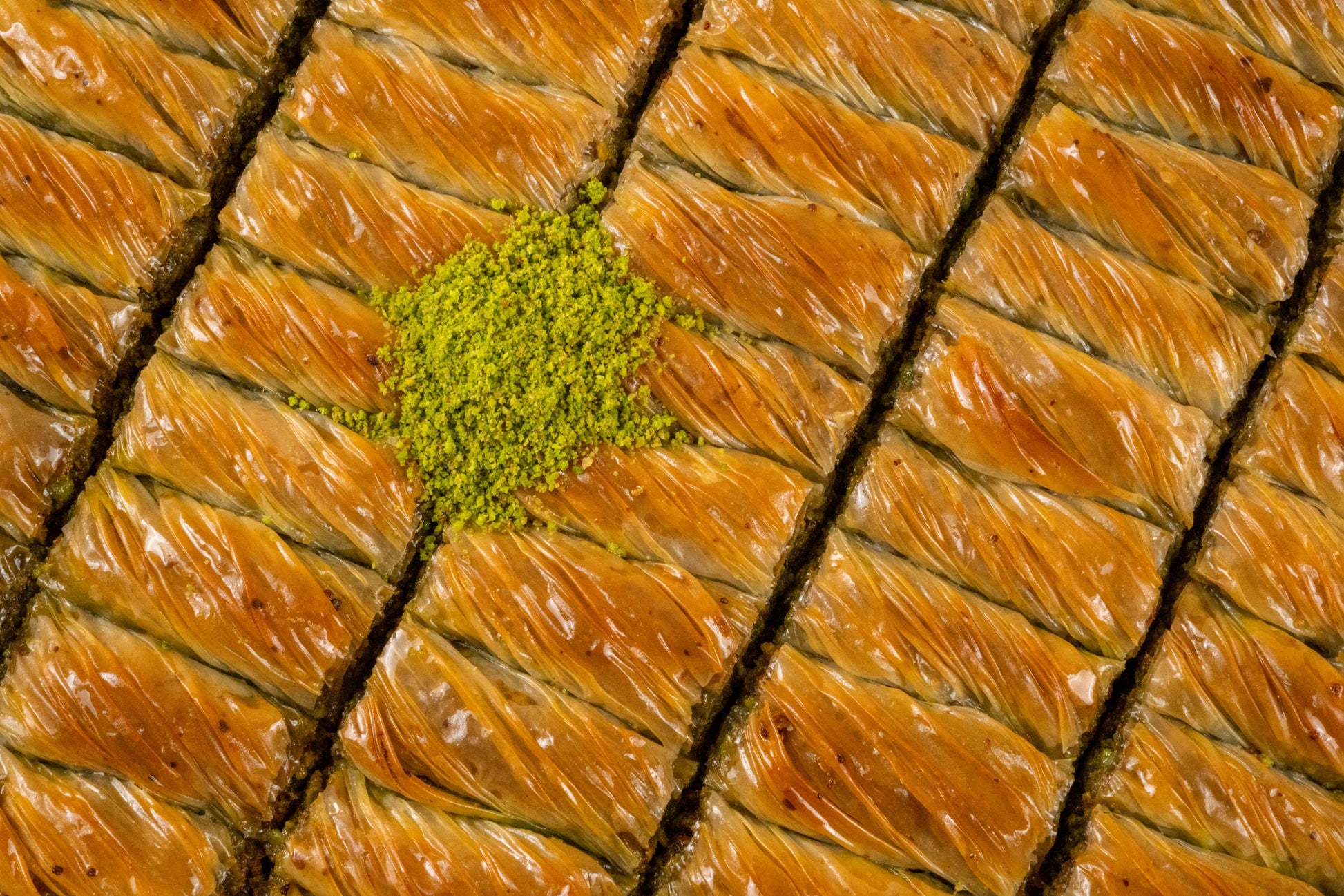 May include: Close-up of a tray of baklava, a traditional Middle Eastern dessert. The golden-brown, layered pastry is cut into square pieces and topped with a vibrant green pistachio crumble. The image highlights the texture and sweetness of the dessert.